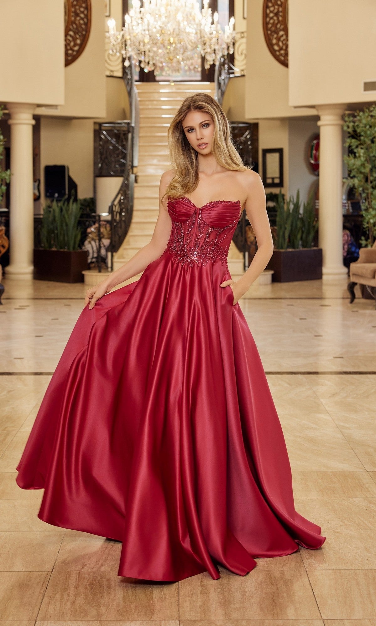 A woman in a burgundy Nox Anabel Y1798 long prom dress with a strapless satin corset bodice and flowing A-line silhouette stands in a grand foyer, with a staircase and chandelier behind her.